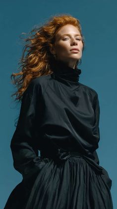 Woman with red hair and freckles wearing a black top and skirt against a blue sky