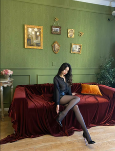 Woman in a sheer black shirt and pantyhose sitting on a red velvet couch