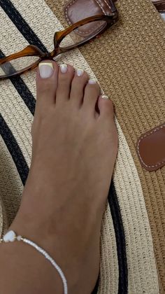 Foot with a white and yellow pedicure, anklet, and reading glasses on a straw bag