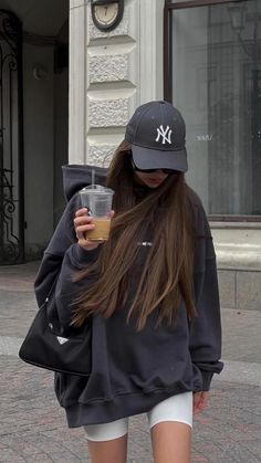 Woman in a dark hoodie and baseball cap holding a drink on a street