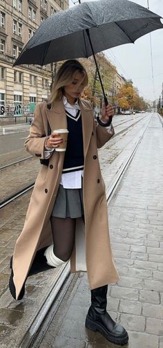 Woman in a long coat and pleated skirt holding an umbrella on a rainy street