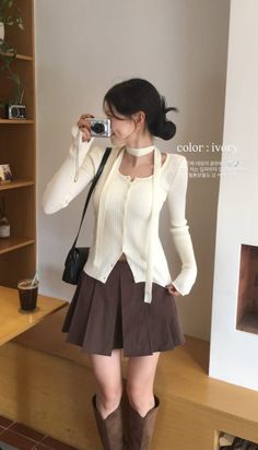 Woman in a cream long sleeve top and brown pleated skirt holding a camera