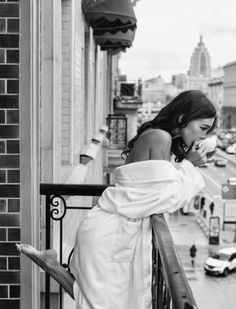 Woman in a white bathrobe drinking from a cup on a balcony overlooking a city