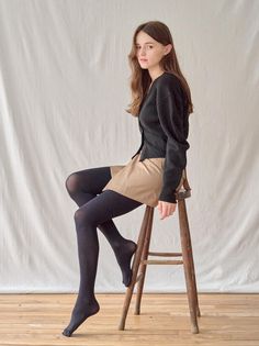 Woman with long hair wearing a black cardigan and tights, sitting on a wooden stool