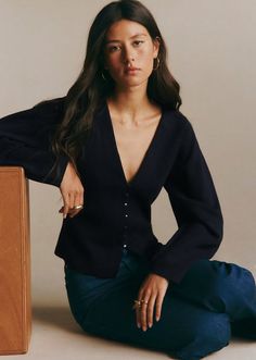 Woman with long dark hair wearing a deep v-neck top and jeans, sitting indoors