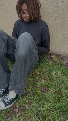 Person with dreadlocks and glasses sitting on grass next to a wall