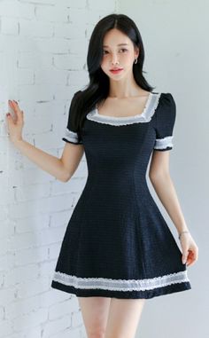 Woman in a black dress with white lace trim posing against a white brick wall