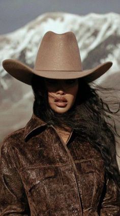 Woman in a cowboy hat and brown jacket with long hair against a mountain background