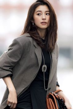 Woman with long brown hair wearing a blazer and long necklace, holding a brown handbag