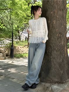 Woman in a white lace top and baggy jeans leaning against a tree outdoors