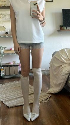 Person in a white t-shirt, lace shorts, knee-high socks, and white heels taking a mirror selfie