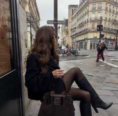 Woman with long hair in a black coat and boots sitting on a city street