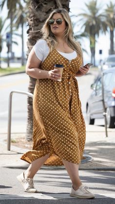 Woman in a polka dot dress and sunglasses walking outdoors with coffee and phone