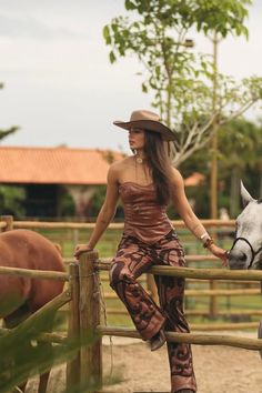 Woman in a cowboy hat and patterned outfit sitting on a fence at a ranch