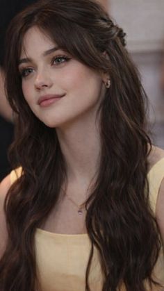 Woman with long wavy brown hair and bangs wearing a yellow top and gold necklace
