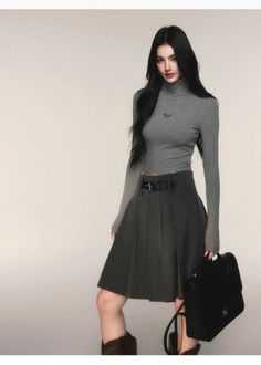 Woman with long black hair wearing a gray turtleneck and pleated skirt, holding a black handbag