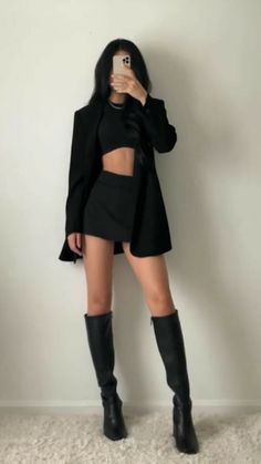 Woman in a black crop top, mini skirt, and long boots taking a mirror selfie