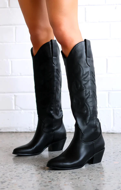 Person wearing black cowboy boots with exposed skin against a white brick wall