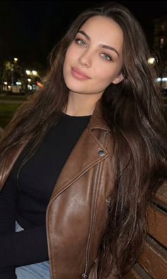 Woman with long brown hair and green eyes wearing a brown leather jacket at night