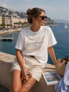 Woman in sunglasses and a white t-shirt sitting by the ocean with a glass of wine