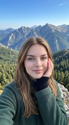 Woman with long hair and green eyes in a green jacket, posing outdoors with mountains and forest in the background