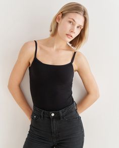Woman with short blonde hair wearing a black camisole and black jeans against a white wall