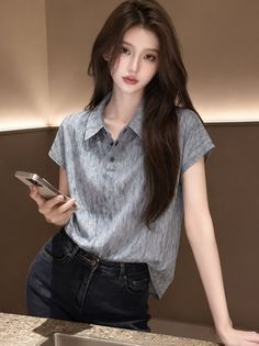 Woman with long hair wearing a polo shirt and jeans, holding a phone indoors
