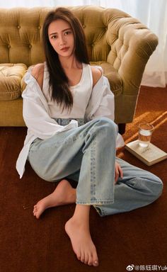 Woman with long hair wearing a white tank top and jeans, sitting on a brown carpet