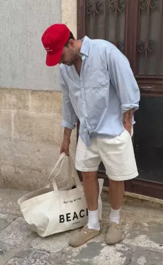 Man in a red cap, light blue shirt, and white shorts holding a tote bag outdoors