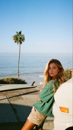 Woman with long blonde hair leaning on a car by the beach with a surfboard