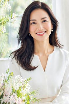 Woman with long dark hair wearing a white top and pearl earrings, smiling indoors