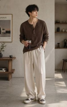 Man in a brown henley shirt and cream sweatpants holding a drink indoors