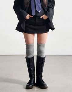 Person in a black skirt, blazer, and knee-high boots with hands in pockets