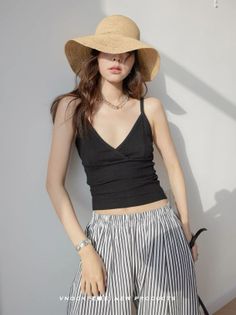 Woman in a straw hat and black top with striped pants, leaning against a wall