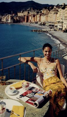 Woman in a floral top and long skirt on a balcony overlooking a seaside town