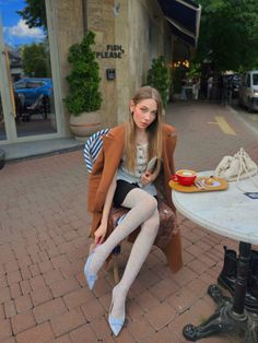 Woman in a brown coat and white tights sitting at an outdoor cafe table