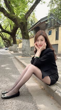 Woman in a black blazer and shorts sitting on a curb on a tree-lined street