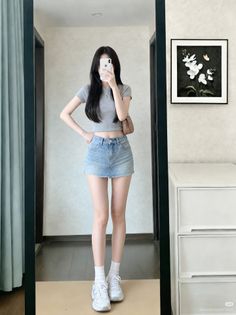 Woman in a crop top and denim skirt taking a mirror selfie indoors