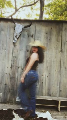 Person in a cowboy hat and jeans posing in front of a wooden fence with a bull skull