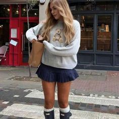 Woman with long blonde hair wearing a grey sweater and pleated skirt on a street