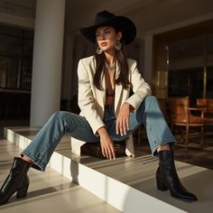 Woman in a cowboy hat, blazer, and jeans sitting indoors with dramatic lighting