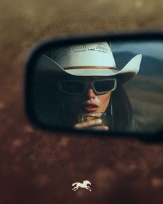 Person in a cowboy hat and sunglasses reflected in a car mirror
