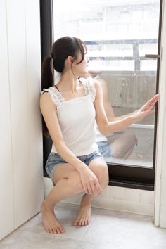 Woman with a ponytail in a white tank top and denim shorts squatting by a window