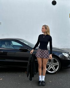 Woman in a plaid skirt and black top standing next to a black car