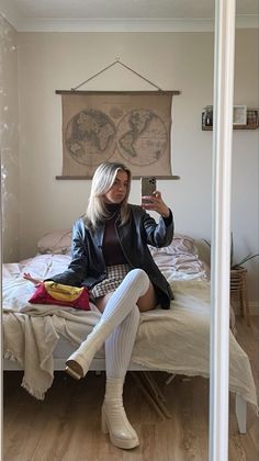 Woman in a leather jacket and plaid skirt taking a mirror selfie in a bedroom