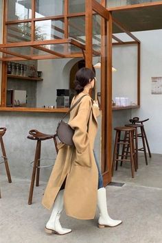 Woman in a long tan coat and white boots walking through a cafe