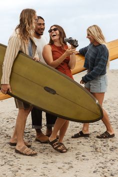 Group of friends with surfboards on a rocky beach, one laughing with a camcorder