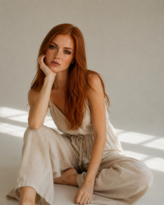 Woman with red hair and freckles wearing a light jumpsuit, sitting on the floor