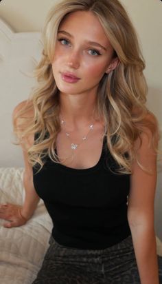 Blonde woman in a black tank top and butterfly necklace, looking at the camera