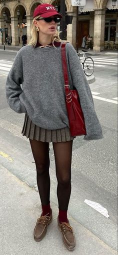 Woman in a grey sweater and pleated skirt with a red baseball cap and bag